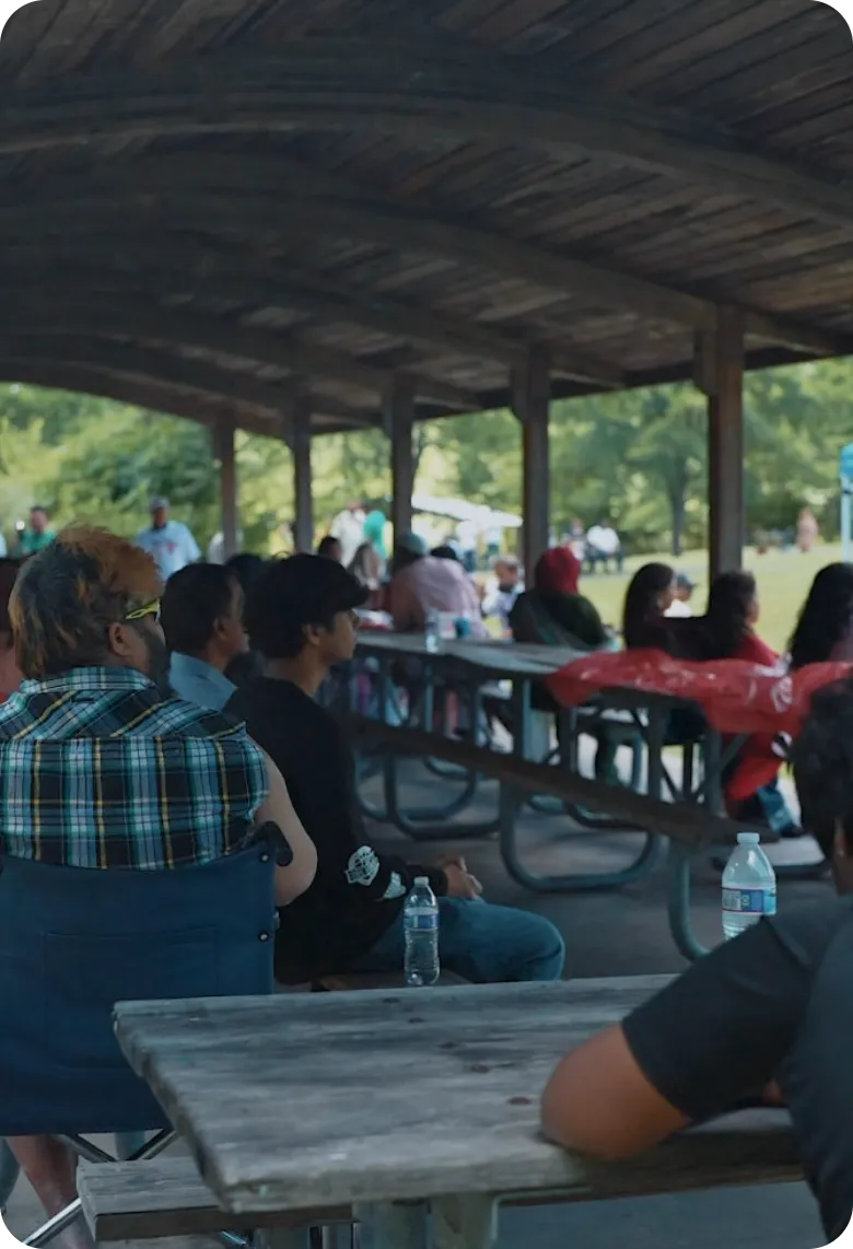 Community event attendees seated at picnic tables under an outdoor park pavilion
